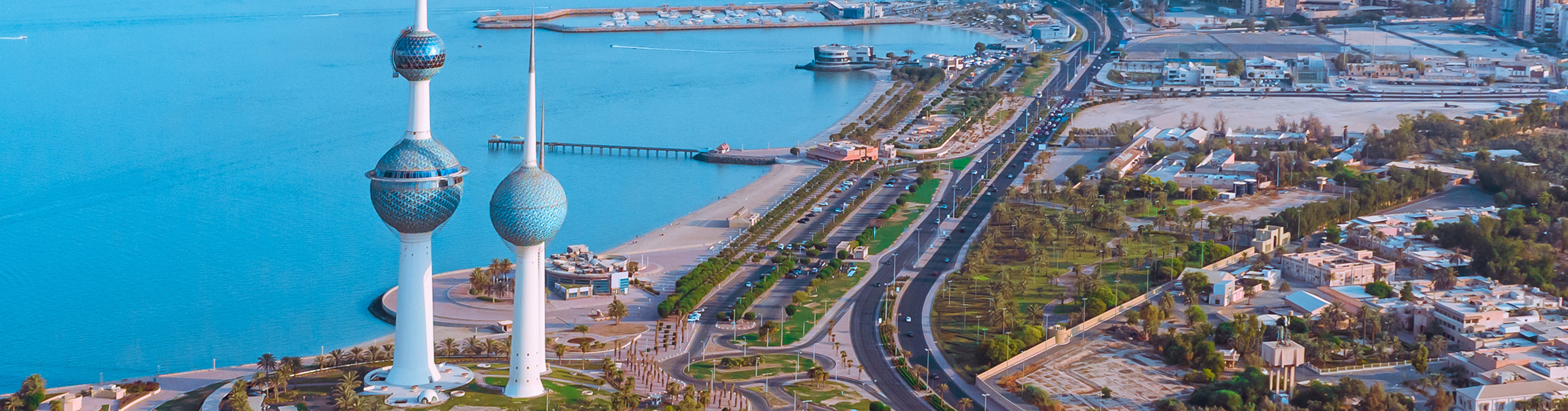 Aerial view of the Kuwait Towers along the Arabian Gulf with nearby roads, greenery, and city buildings.